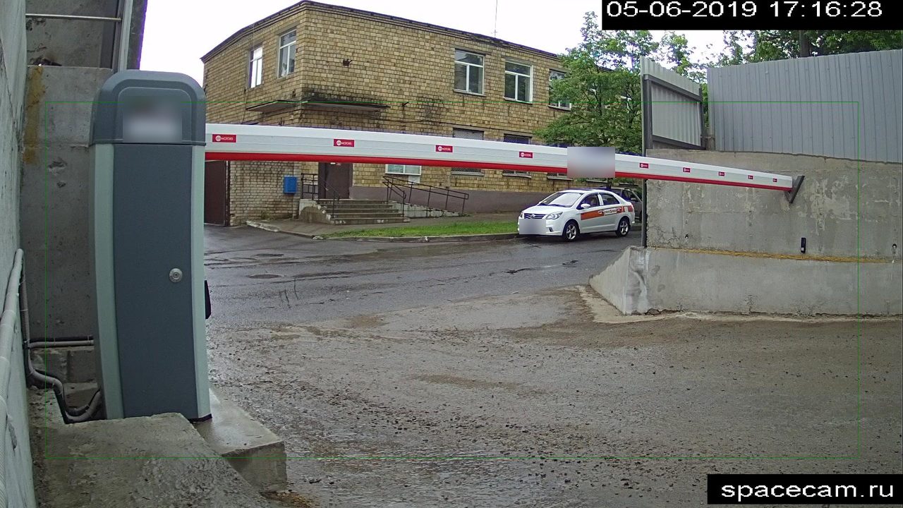 A camera frame at the barrier: the lowered boom, the drive cabinet, a courtyard and a taxi behind